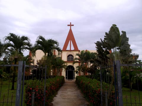 La iglesia de San Rafael de Guatuso al final de un camino con jardines de flores rojas y palmeras.