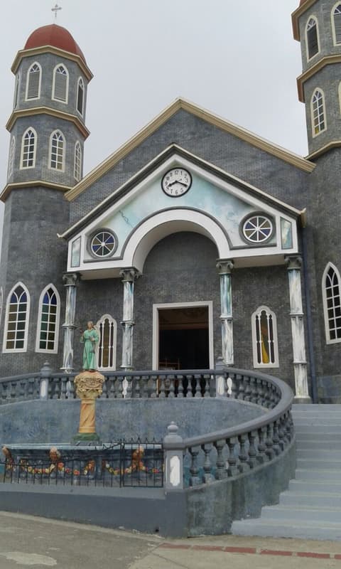 Fachada de la iglesia de Zarcero, de tonos grises y celestes, con dos torres de cúpula roja y un reloj sobre la entrada.