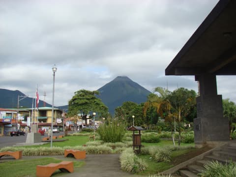 Jardines y bancas del parque de La Fortuna con el cono del volcán Arenal detrás de los edificios del centro.